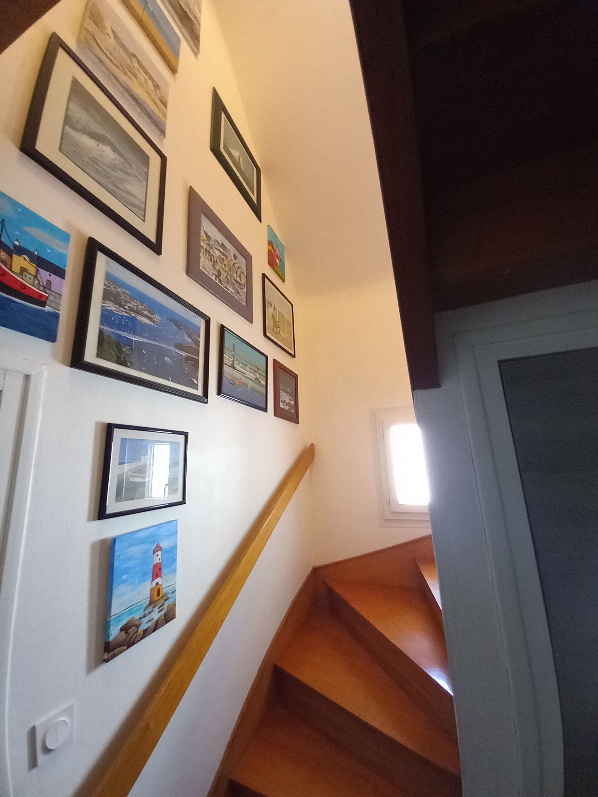 Un escalier avec des marches en bois et une rampe, menant à un palier dont le mur blanc est orné d’œuvres d’art encadrées.