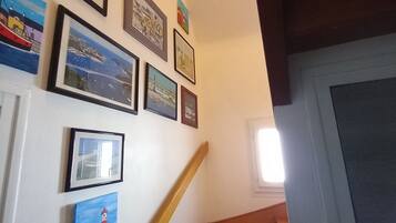 Un escalier avec des marches en bois et une rampe, menant à un palier dont le mur blanc est orné d’œuvres d’art encadrées.