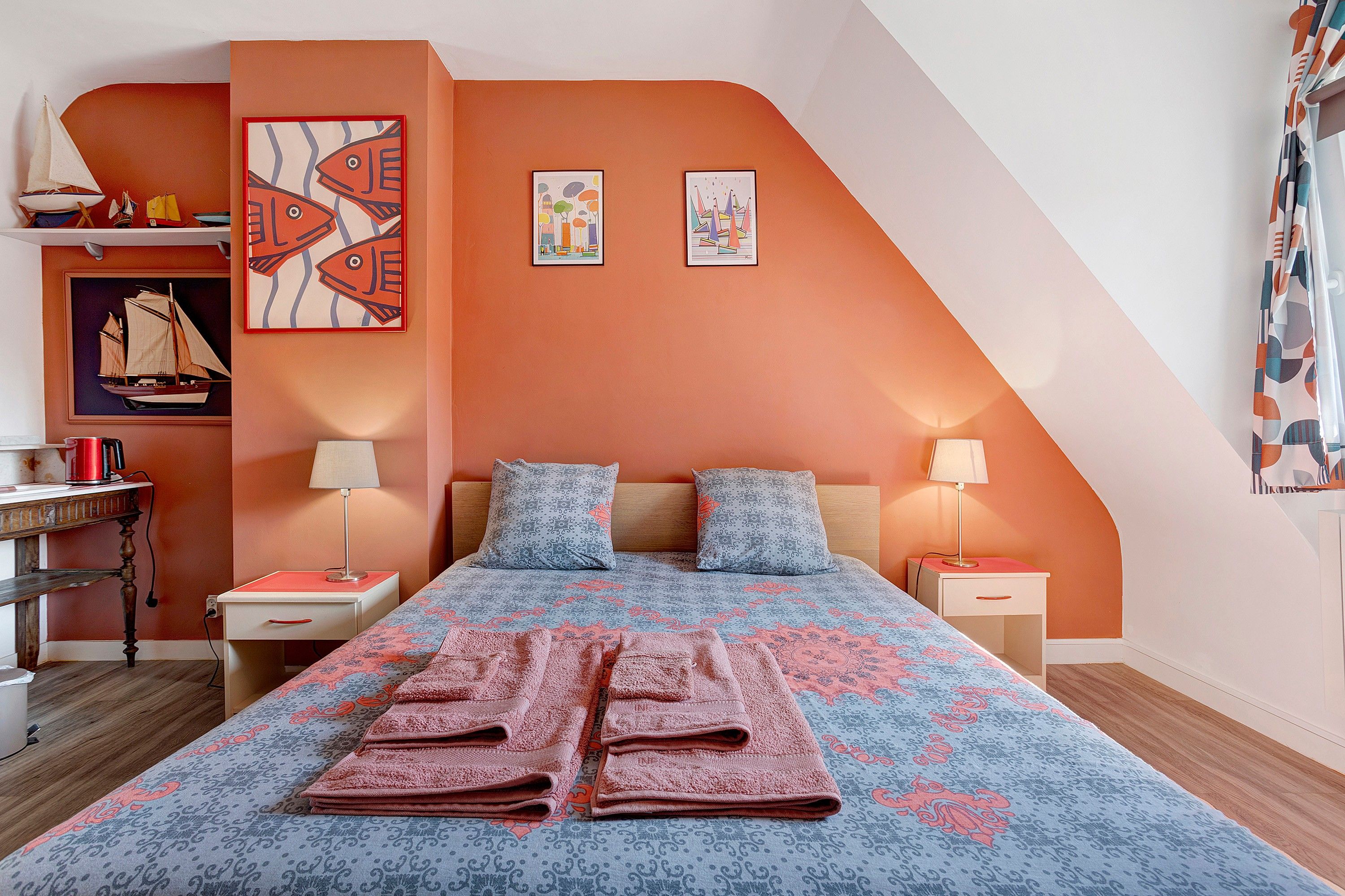 Une chambre avec un plafond en pente, un lit avec une literie à motifs, deux oreillers, des tables de chevet avec des lampes et des œuvres d’art accrochées aux murs.