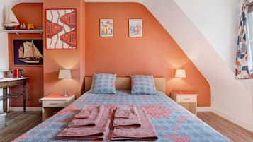 Une chambre avec un plafond en pente, un lit avec une literie à motifs, deux oreillers, des tables de chevet avec des lampes et des œuvres d’art accrochées aux murs.