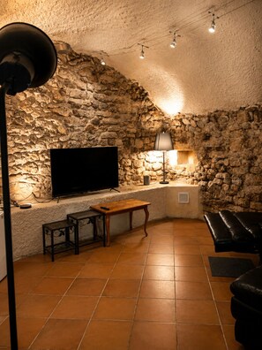 A room with a stone wall, a flat-screen TV, a wooden bench, a table, and a floor lamp. - Former Loft Studio, 105 m² • Air Conditioning • Vaulted Cellar • The Heart of Aix's Historic Center (Aix-en-Provence)