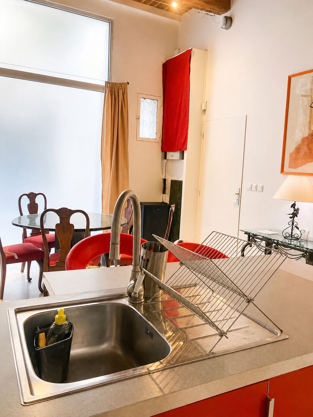 A modern kitchen with a stainless steel sink, a drying rack, and a dining area with red chairs.