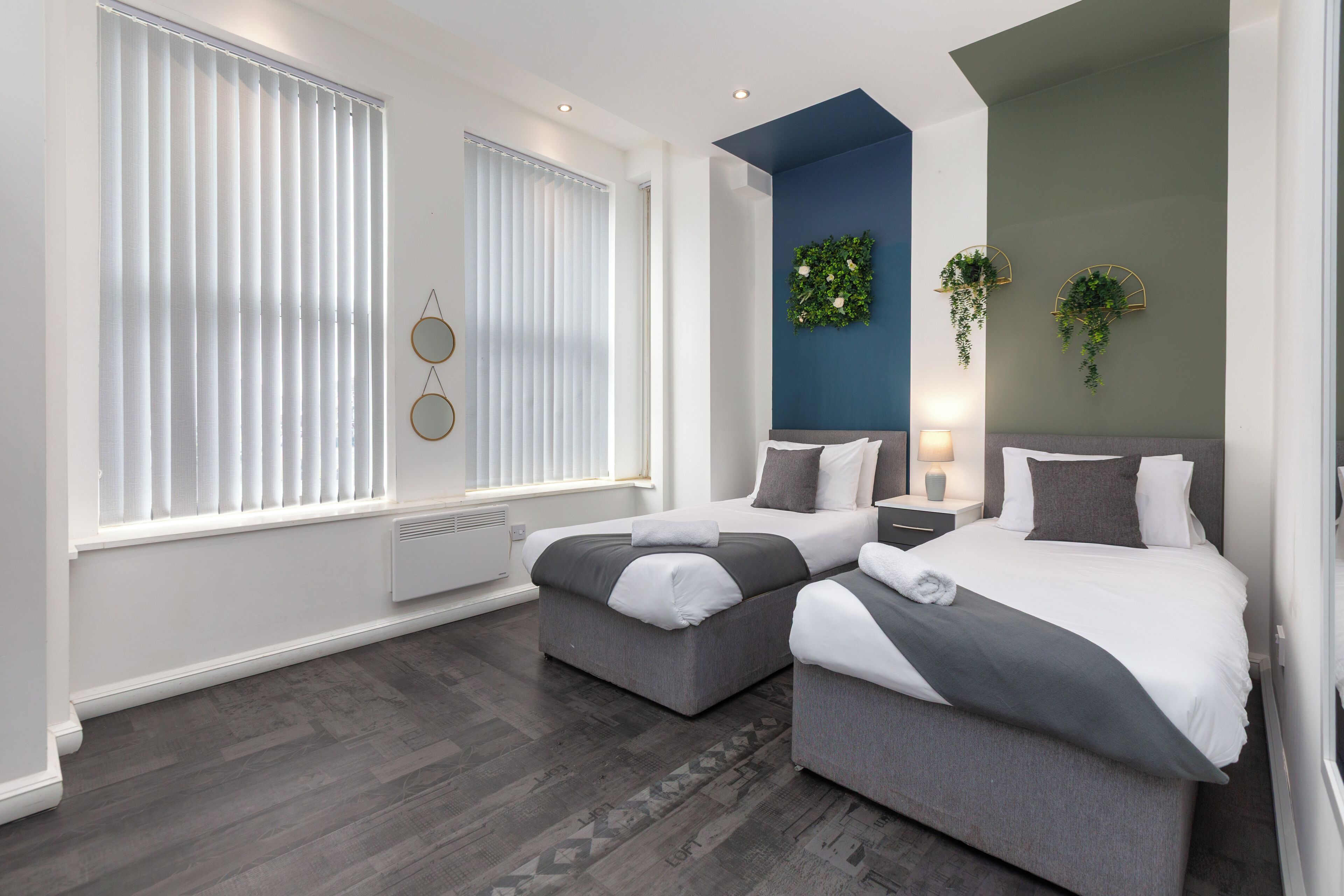 Une chambre moderne avec deux lits, un radiateur, une fenêtre avec des stores et une plante fixée au mur.