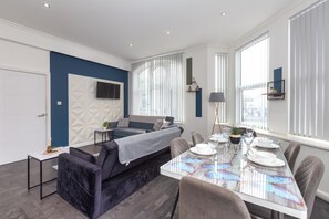A modern living space with a dining area, a sofa, a TV mounted on the wall, and a window with blinds. - South Pier Apartment 4 (Blackpool)