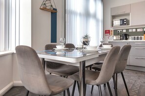 A modern dining area with a table set for four, surrounded by chairs, in a well-lit room with a kitchen in the background. - South Pier Apartment 4 (Blackpool)