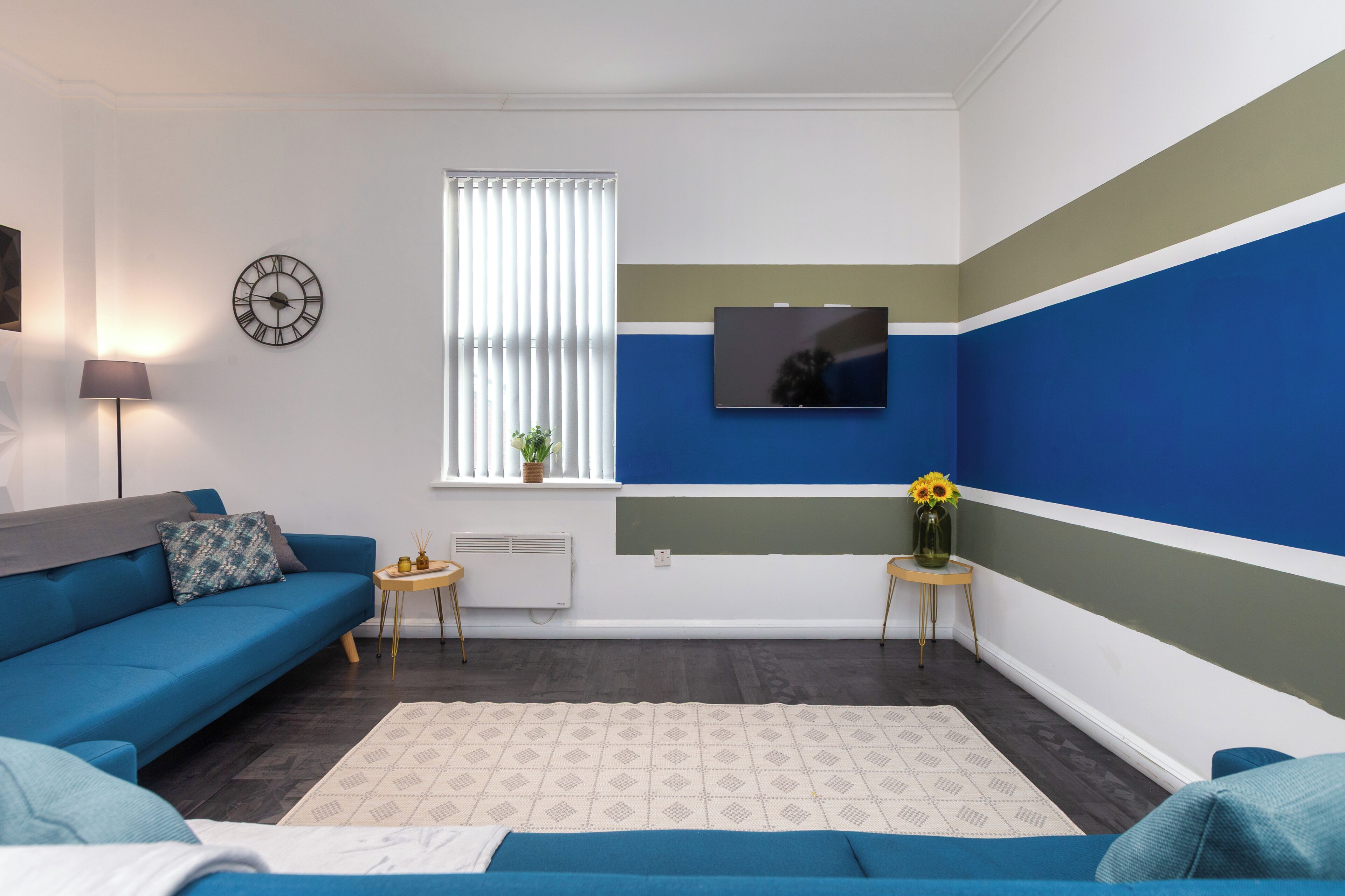 Una sala de estar moderna con un sofá azul, una mesita auxiliar, una lámpara de pie, un televisor montado en la pared y una ventana con persianas.