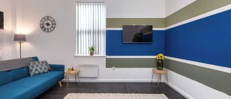 Una sala de estar moderna con un sofá azul, una mesita auxiliar, una lámpara de pie, un televisor montado en la pared y una ventana con persianas.