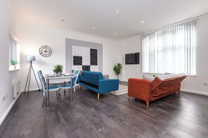 A modern living room with a dining area, a blue sofa, an orange sofa, a TV mounted on the wall, and a large window with blinds. - South Pier Apartment 6 (Blackpool)