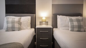 Ein Hotelzimmer mit zwei Einzelbetten, jeweils mit Kissen und Nachttisch mit Lampe. Zwischen den Betten steht eine kleine Kommode.