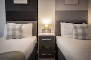 Ein Hotelzimmer mit zwei Einzelbetten, jeweils mit Kissen und Nachttisch mit Lampe. Zwischen den Betten steht eine kleine Kommode.