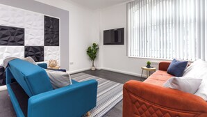 Ein modernes Wohnzimmer mit einem blau-orangen Sofagarnitur, einem an der Wand montierten Flachbildfernseher und einer Wand mit geometrischem Muster.