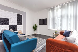 Ein modernes Wohnzimmer mit einem blau-orangen Sofagarnitur, einem an der Wand montierten Flachbildfernseher und einer Wand mit geometrischem Muster.