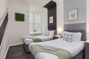 A hotel room with two beds, a window with blinds, a wall-mounted plant, and a framed picture. - South Pier Apartment 6 (Blackpool)