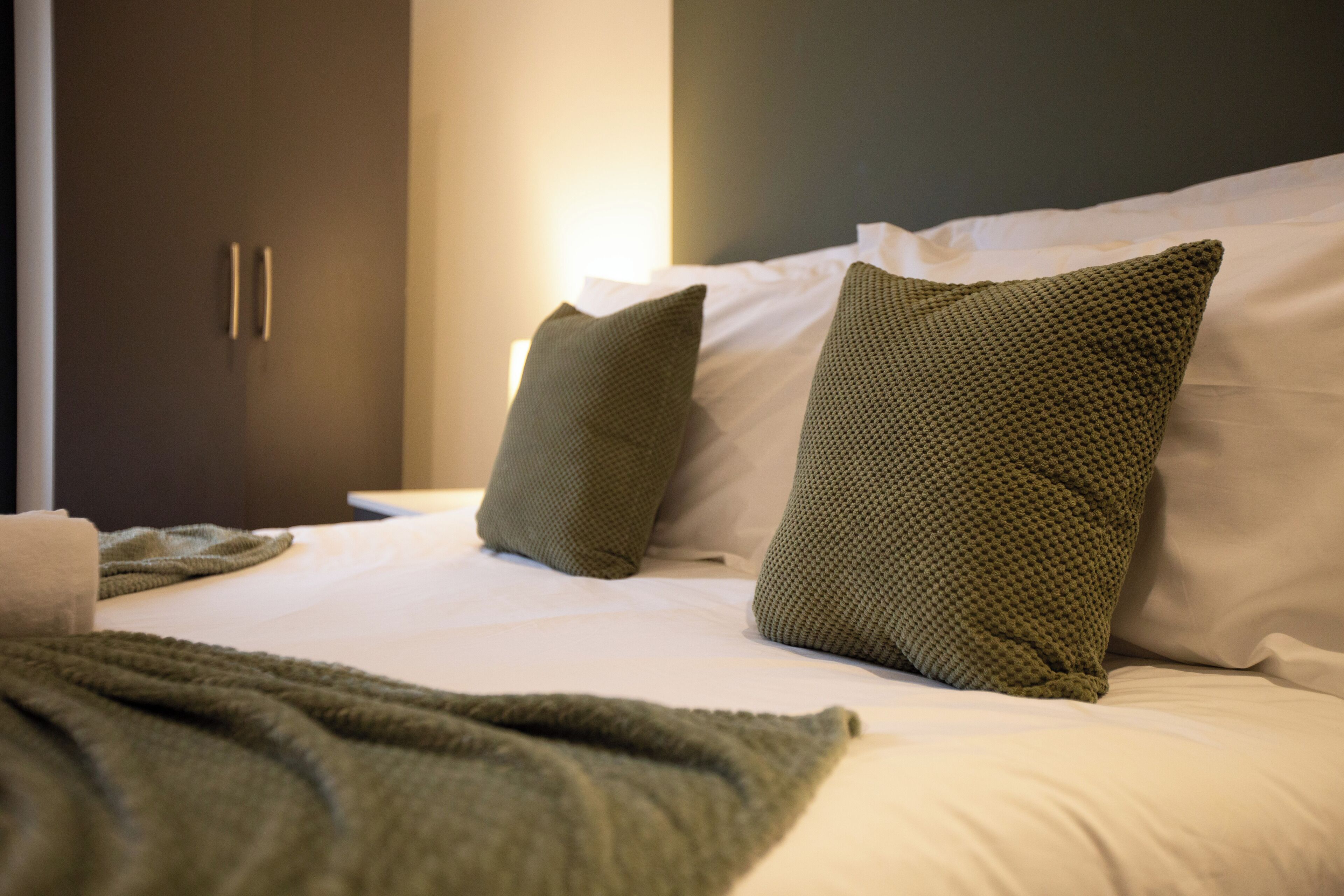 A neatly made bed with two pillows and a blanket, a dark-colored wardrobe, and a white wall.