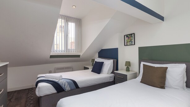 A bedroom with two beds, a window, a radiator, and a painting on the wall. - South Pier Apartment 7 (Blackpool)