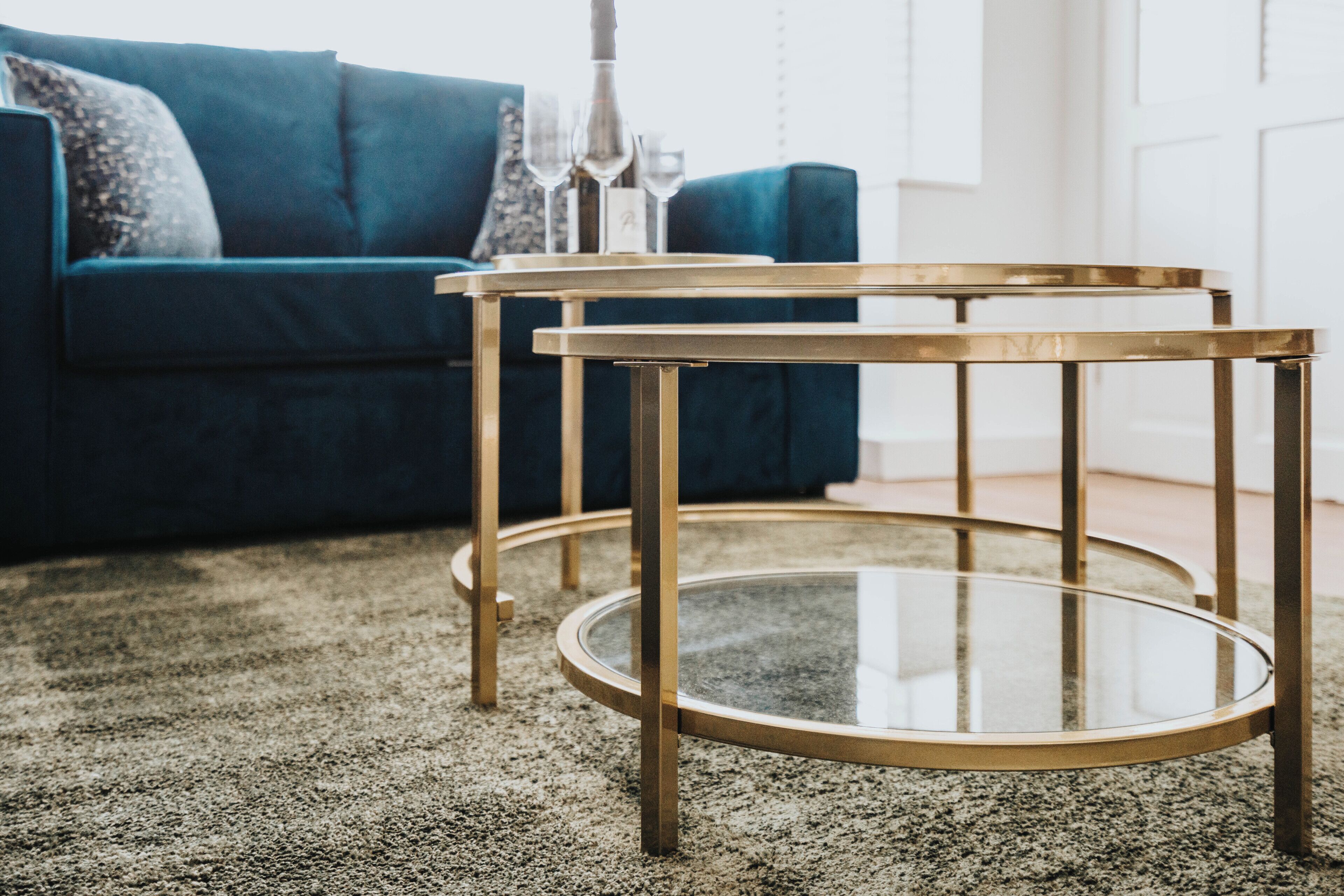A room with a blue sofa, two gold-framed glass tables, and a carpeted floor.