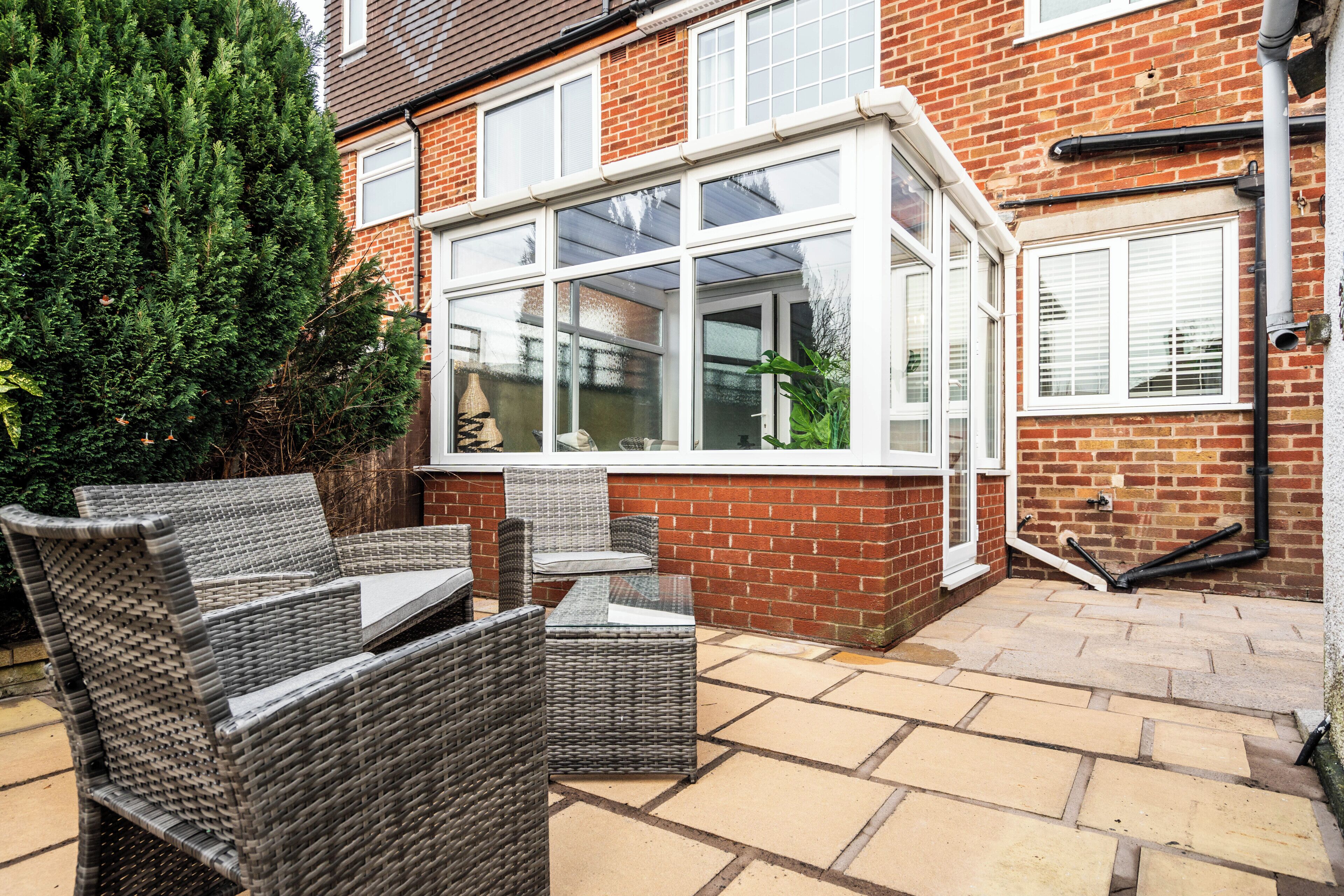 A patio area with wicker furniture and a glass table, adjacent to a brick house with a white-framed window.
