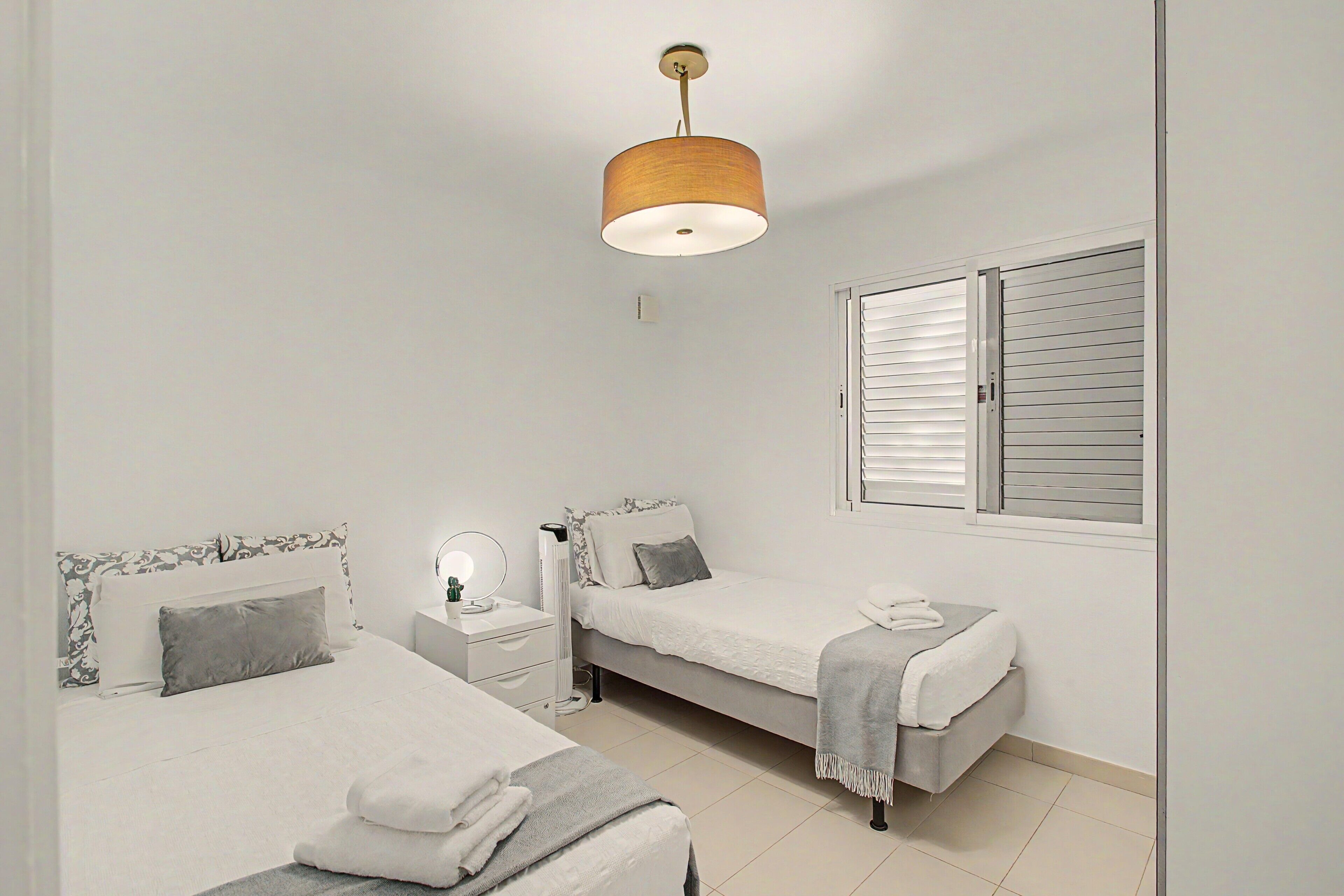 Une chambre avec deux lits, une table de chevet avec une lampe, une fenêtre avec des volets et un luminaire suspendu.
