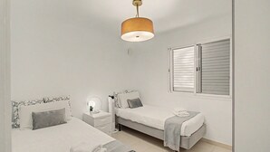 A bedroom with two beds, a bedside table with a lamp, a window with shutters, and a hanging light fixture. - Nueva Suecia 805 – Beachfront Apartment with Terrace and Sea Views (San Bartolomé de Tirajana)