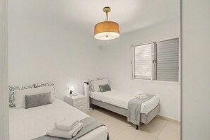 Quarto com duas camas, mesa de cabeceira com abajur, janela com persianas e luminária pendente.