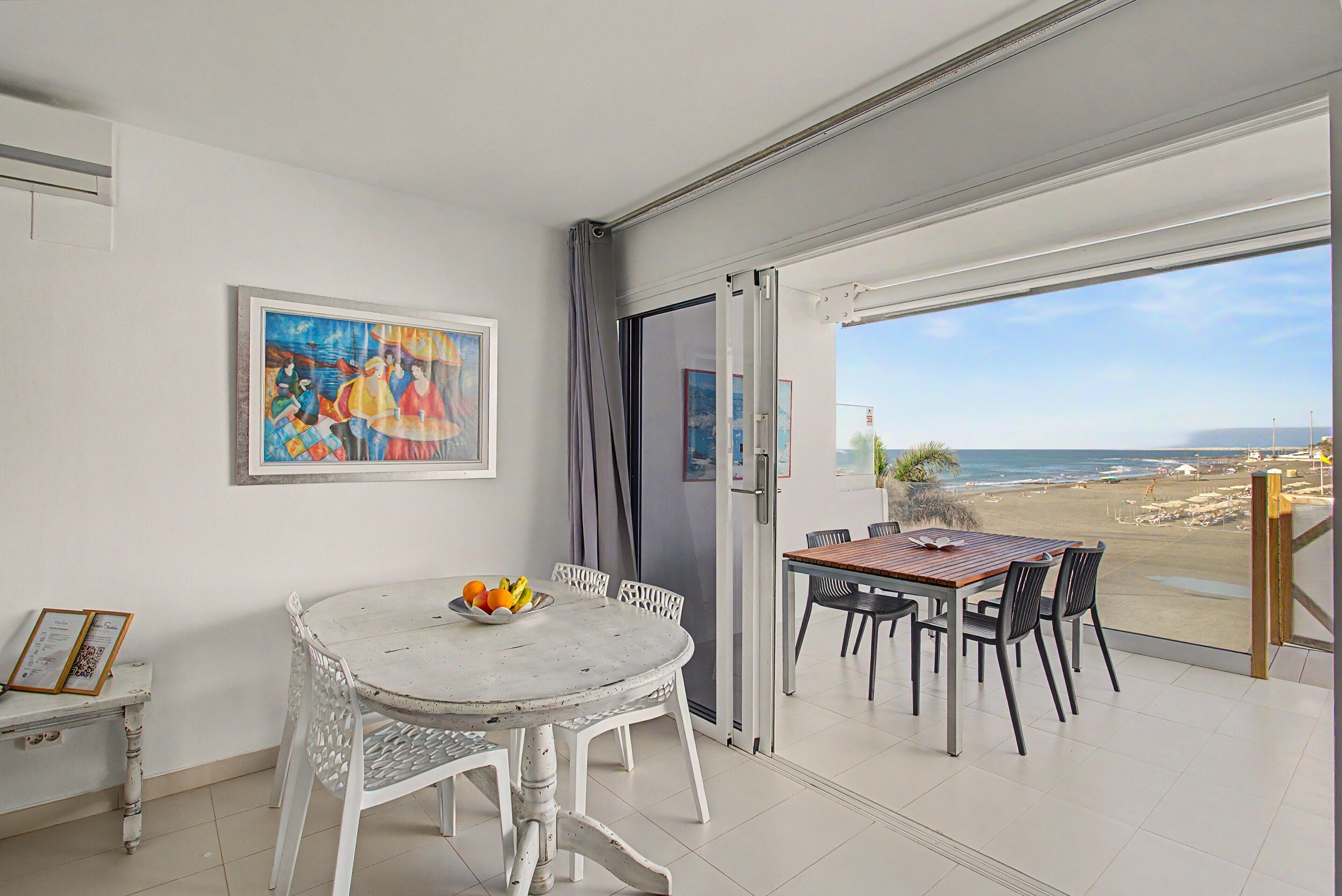Une pièce comprenant une table à manger et des chaises, un tableau accroché au mur, et une vue sur la plage par une porte ouverte.