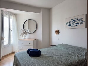 Ein Schlafzimmer mit einem Bett, einer Kommode, einem Spiegel, einem an der Wand befestigten Ventilator und einem Kunstwerk mit Fischmotiven.