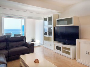 A modern living room with a leather sofa, a flat-screen TV on a white entertainment unit, and a view of the ocean through a large window. - Apartment Clara by Interhome (St Antoni de Calonge)