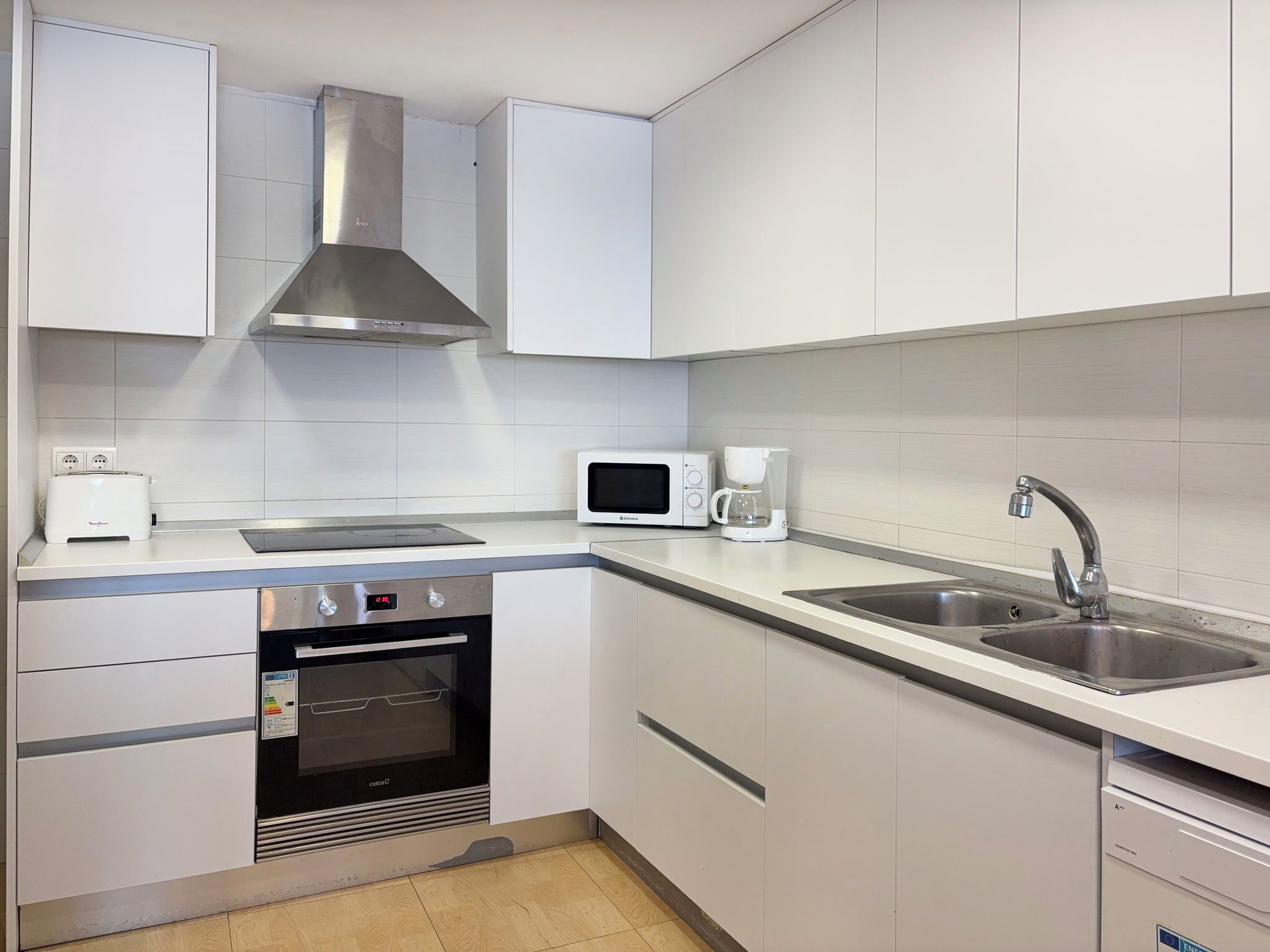 Una cocina moderna con gabinetes blancos, una campana extractora de acero inoxidable, microondas, cafetera, fregadero y horno.