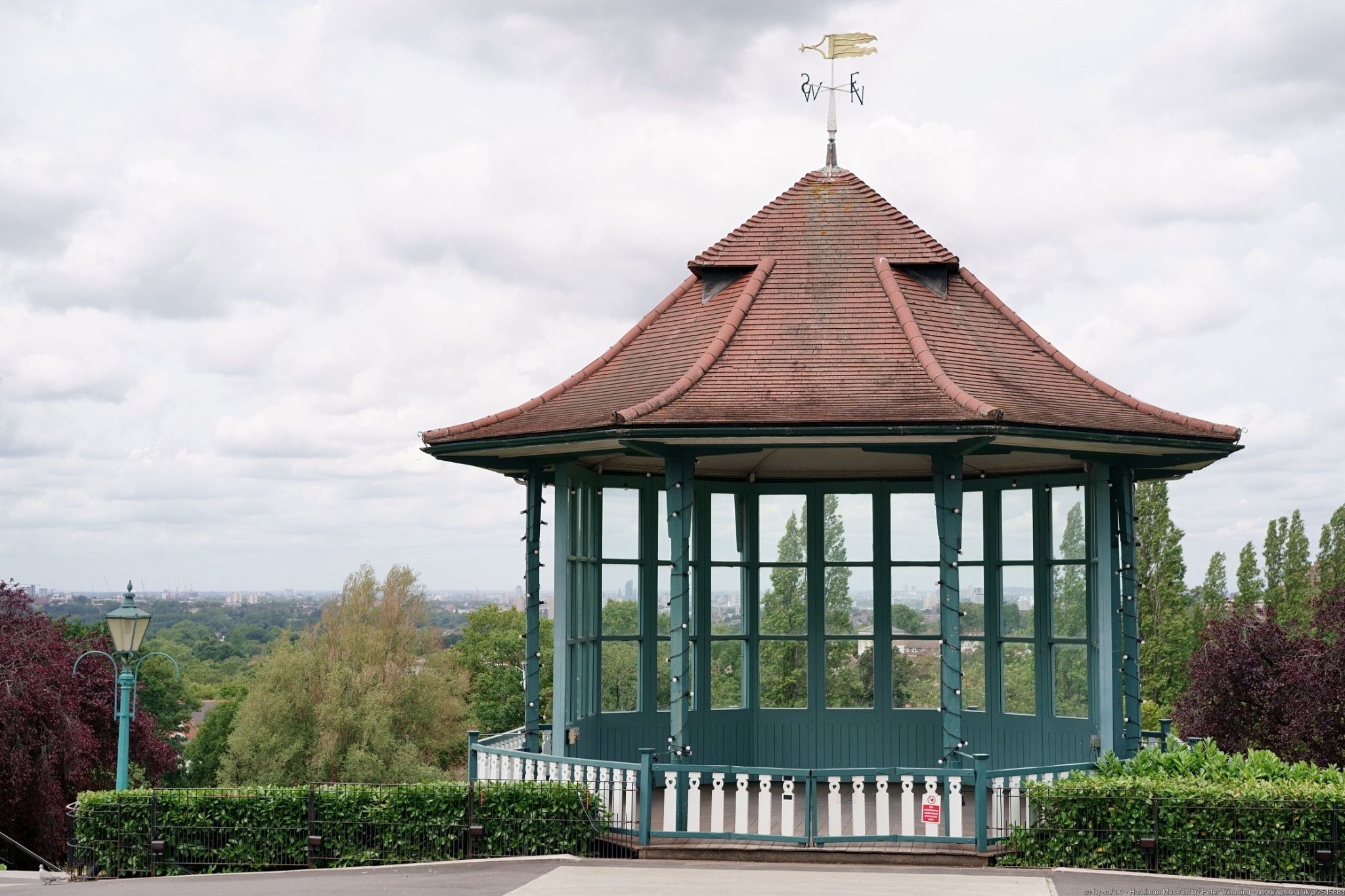 A gazebo with a peaked roof and a weather vane, surrounded by greenery and a cityscape in the background.