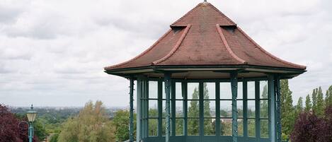A gazebo with a peaked roof and a weather vane, surrounded by greenery and a cityscape in the background.