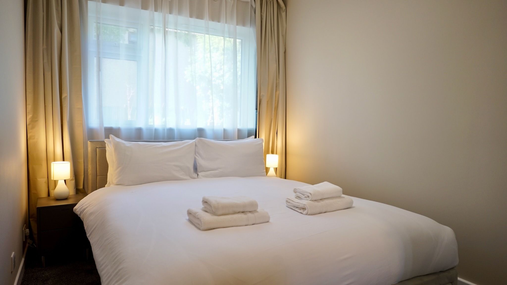 A hotel room with a large bed, two pillows, a nightstand with a lamp, and a window with curtains.