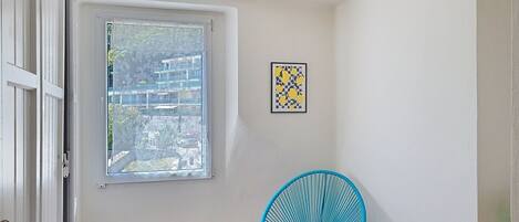 A room with a window, a wall-mounted light fixture, a framed artwork, and a blue chair.