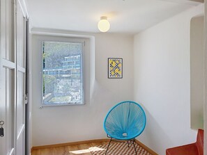 A room with a window, a wall-mounted light fixture, a framed artwork, and a blue chair. - Casa Yakamos by Interhome (Brissago)