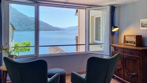 A room with a large window offering a view of a lake and mountains, two chairs, a small table with a plant, and a wooden cabinet with a radio. - Casa Yakamos by Interhome (Brissago)