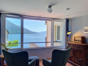 A room with a large window offering a view of a lake and mountains, two chairs, a small table with a plant, and a wooden cabinet with a radio. - Casa Yakamos by Interhome (Brissago)
