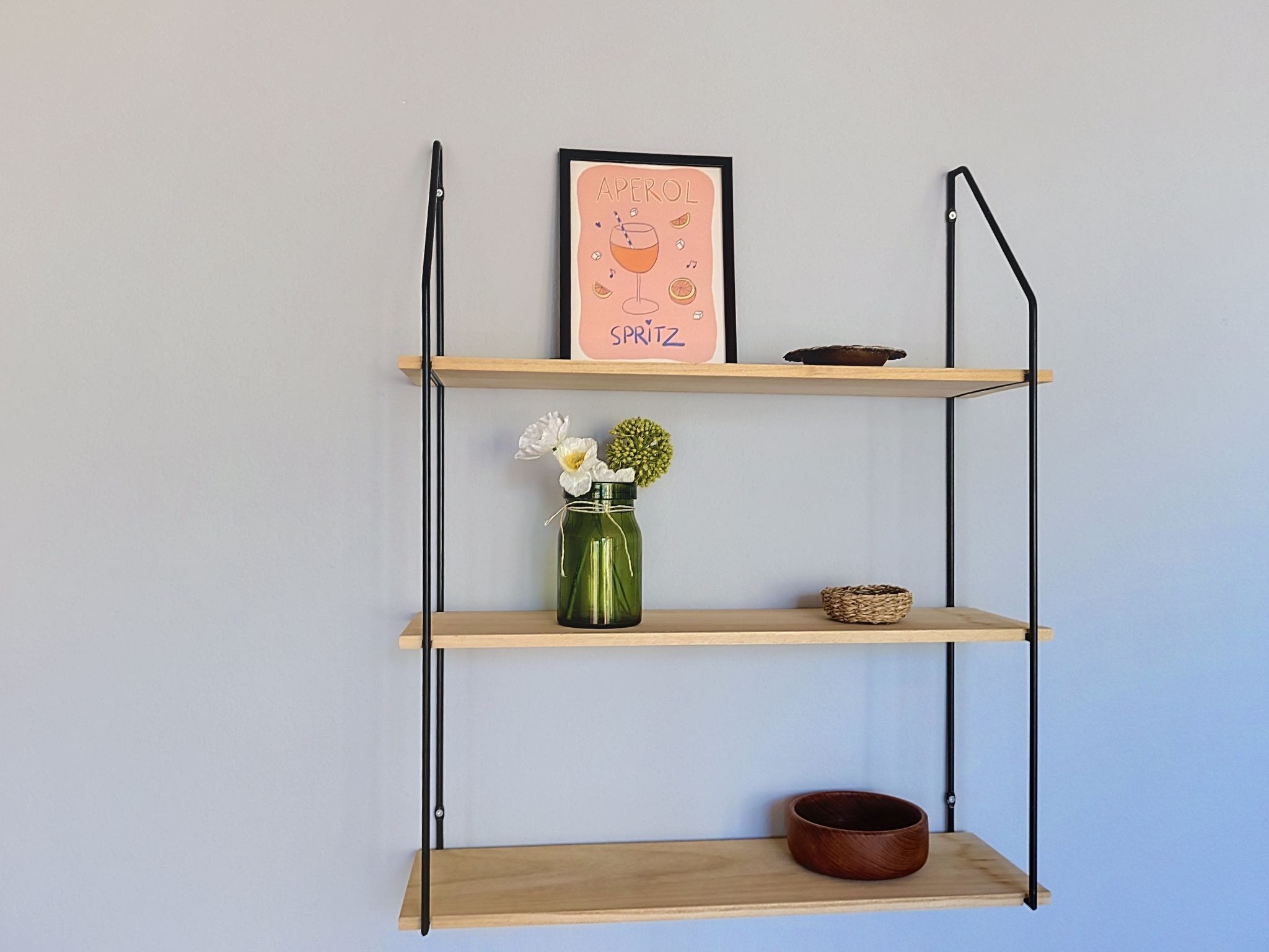 Un estante de pared con tres repisas, un cuadro enmarco, un florero verde con flores y un tazón marrón.