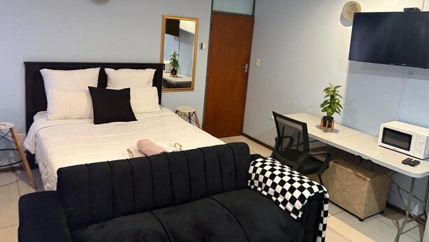 Un dormitorio con cama, escritorio, silla, sofá negro, manta a cuadros, espejo y televisión.