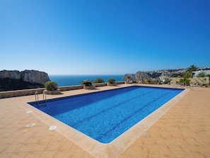 A rectangular swimming pool with clear blue water, surrounded by a tiled deck, set against a backdrop of a coastal landscape with cliffs and a clear sky. - Pueblo de la Luz by Interhome (Moraira)
