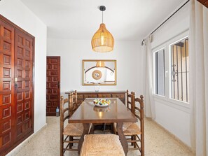 Une salle à manger avec une table en bois, des chaises, un luminaire suspendu, un miroir et une fenêtre avec des barreaux.