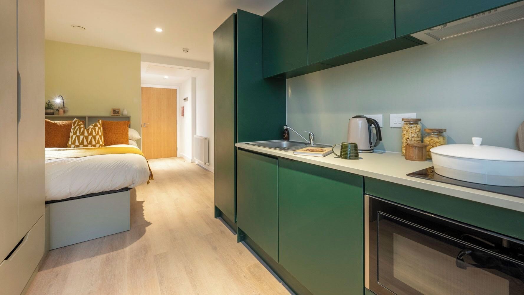 A compact hotel room with a kitchenette, a bed with orange bedding, and a wooden door.