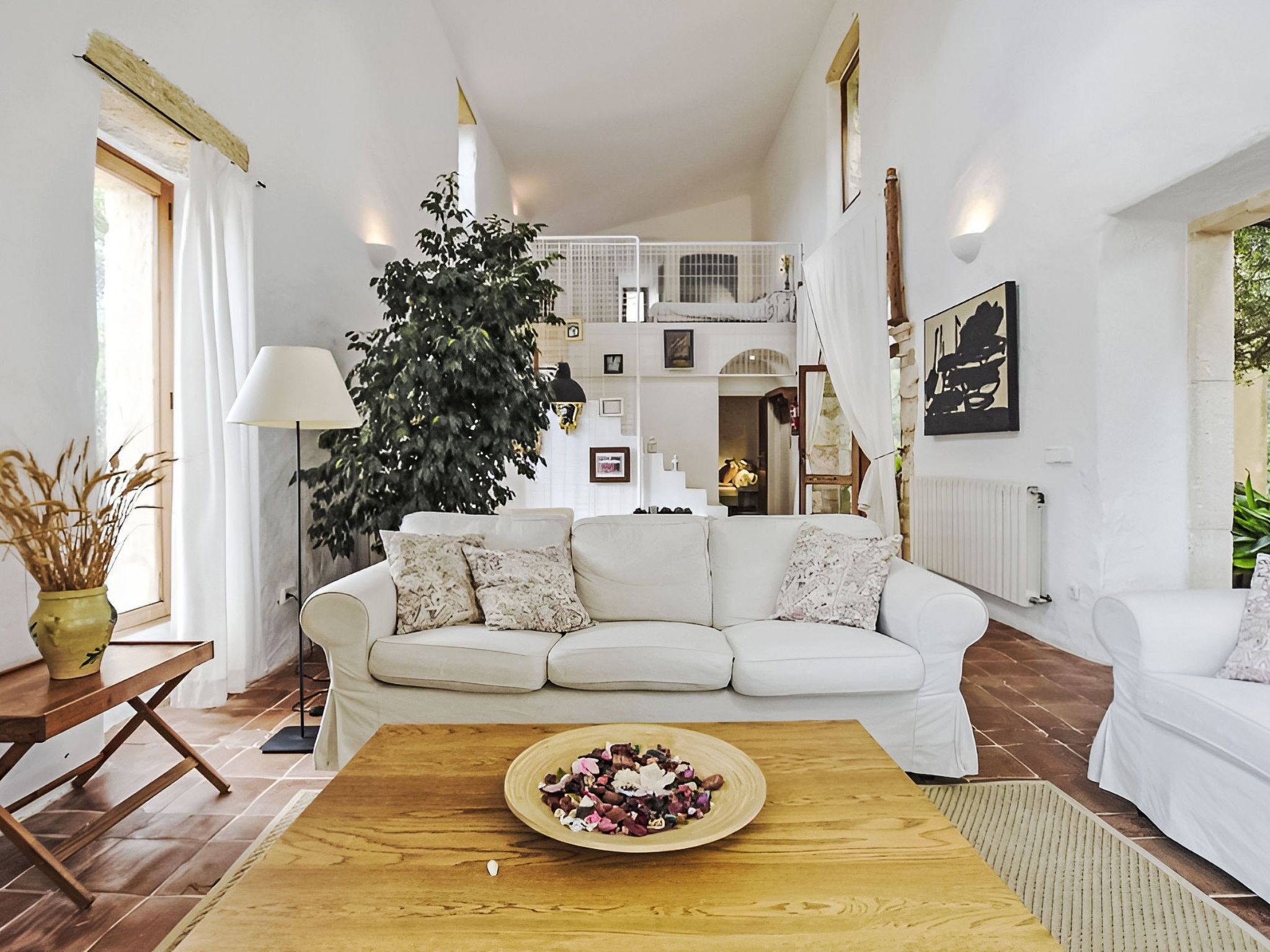 Un salon avec un canapé blanc, une table basse sur laquelle est posé un vase de fleurs, une grande plante et un escalier menant à l’étage supérieur.