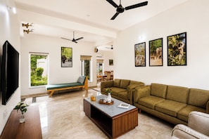 Ein geräumiges Wohnzimmer mit einem grünen Sofa, einem Couchtisch und einem Fernseher. Es gibt Deckenventilatoren, einen Essbereich und ein Fenster mit Blick auf Grünflächen.