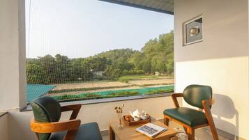 Quarto com uma grande janela com vista para uma piscina e vegetação, duas cadeiras, uma mesa pequena com uma bandeja de objetos e um quadro na parede.