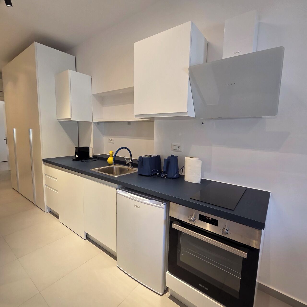 Una cocina moderna con armarios blancos, un refrigerador de acero inoxidable, lavavajillas, horno empotrado y encimera negra.