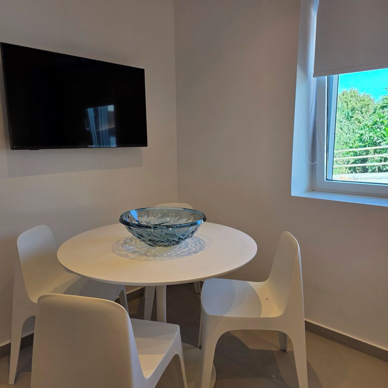 Un espace repas moderne avec une table ronde, un centre de table en forme de bol en verre et des chaises blanches. Un téléviseur à écran plat est fixé au mur, au-dessus de la table.