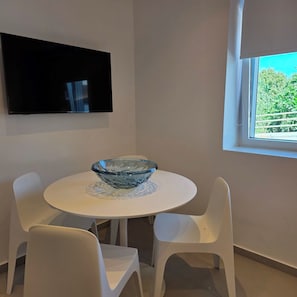 Een moderne eetruimte met een ronde tafel, een glazen schaal als middelpunt en witte stoelen. Een flatscreen televisie is aan de muur boven de tafel bevestigd.