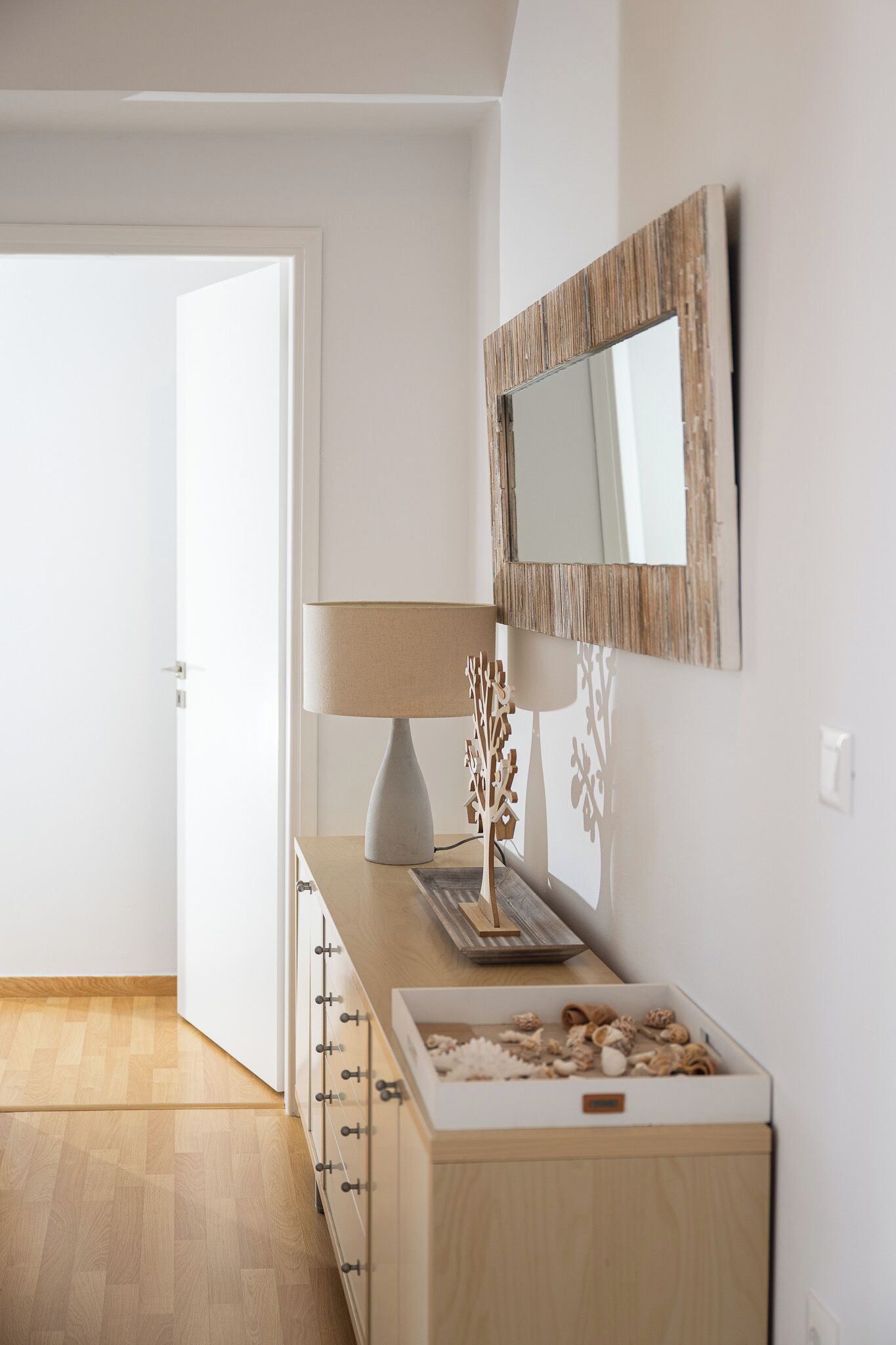 Quarto minimalista com cômoda de madeira, abajur, objeto decorativo e espelho na parede.