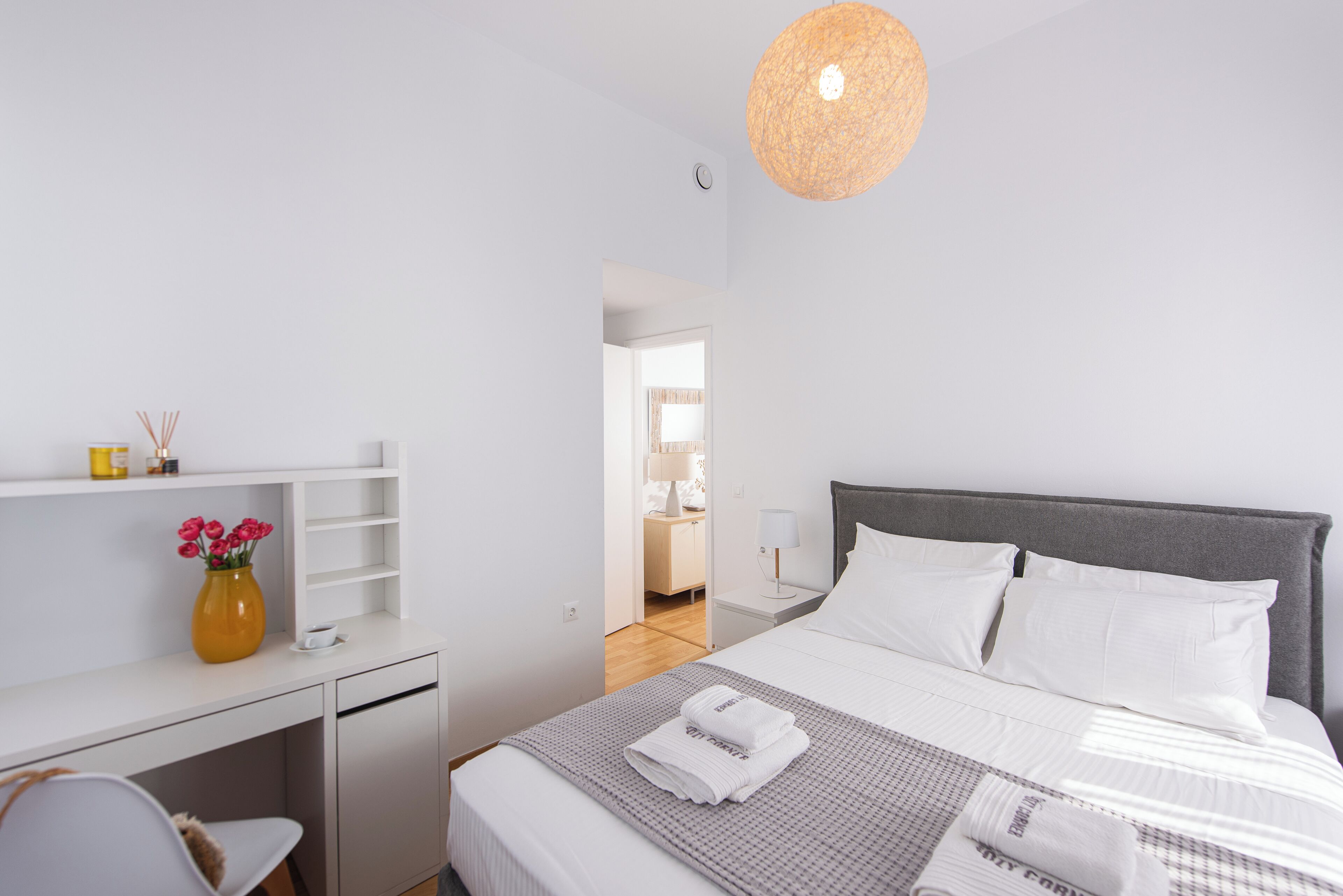 A modern bedroom with a bed, bedside table, and a desk with a vase of flowers.
