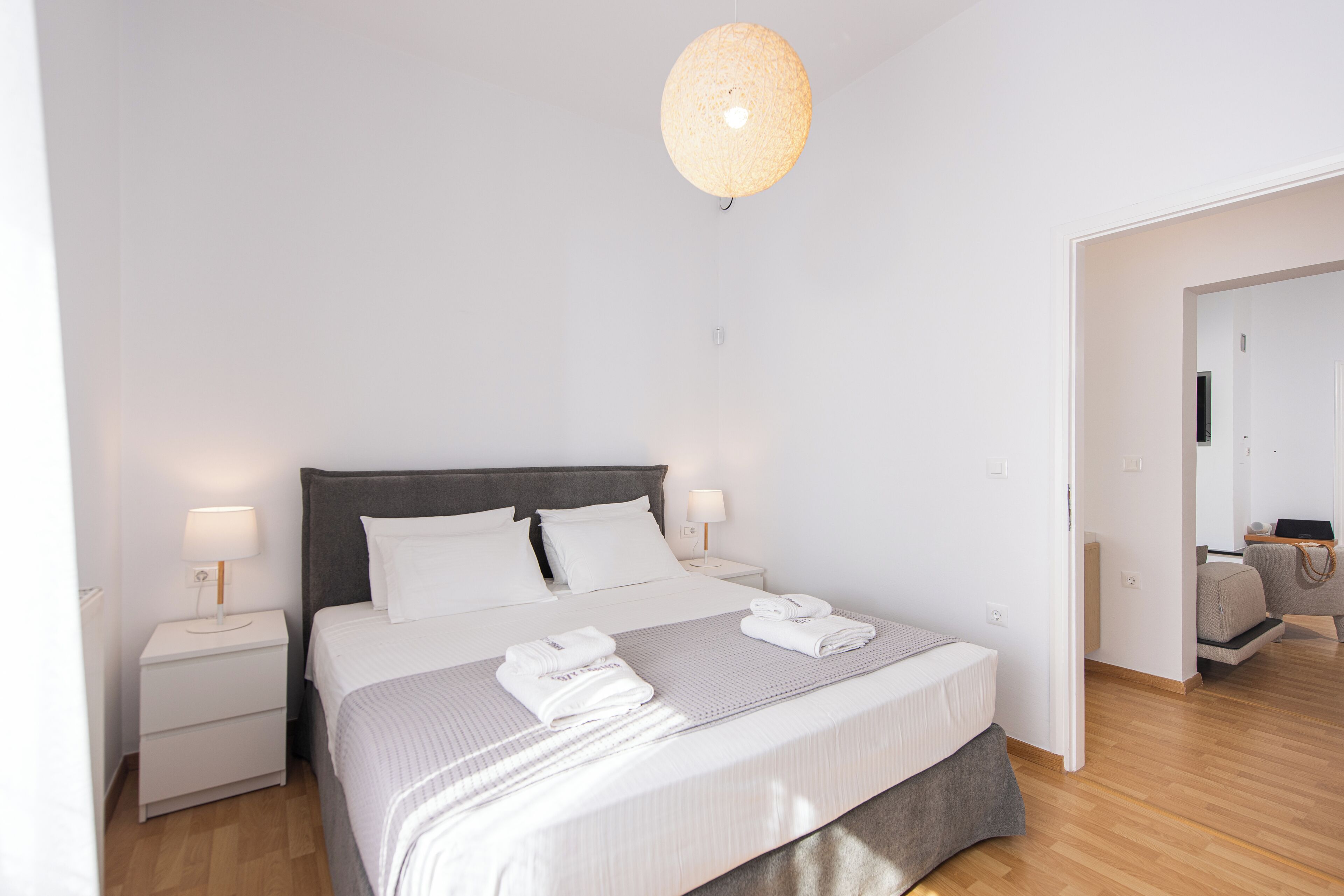 A modern bedroom with a large bed, two bedside tables, a hanging light fixture, and a view into an adjacent room.