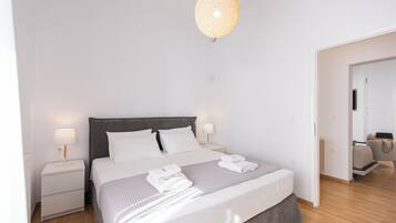 A modern bedroom with a large bed, two bedside tables, a hanging light fixture, and a view into an adjacent room.